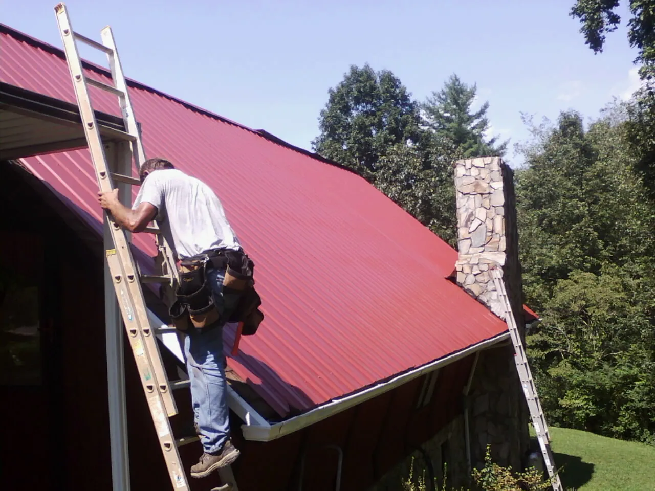 Roofer on a ladder beside a red metal roof and stone chimney — Soffit & Fascia Repair by Roofing Brothers in Lamont